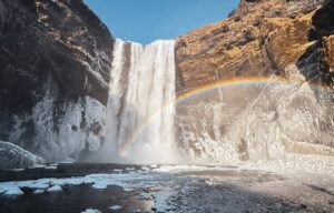 Skógafoss waterfall plunges dramatically over a steep cliff, surrounded by icy rock formations. A vibrant rainbow arcs through the mist, adding a magical touch to the powerful cascade. The dark, rugged cliffs contrast with patches of golden grass, while chunks of ice float on the river below, highlighting the wintery conditions. Bright sunlight and a clear blue sky complete the breathtaking scene.