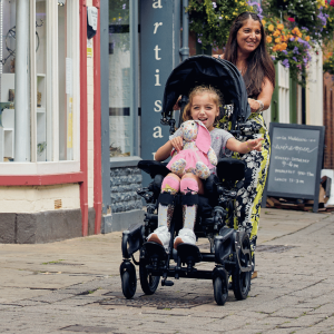 Lilac in her wheelchair with raincover being pushed by her mum down a street in Hereford, both smiling at the camera