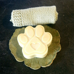 A cream grass-fed tallow paw print dog soap with a heart detail, resting on a decorative leaf dish, alongside a natural knitted wash cloth, handmade by Fatglow in Taranaki New Zealand