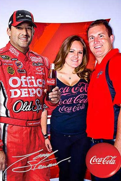 A Coca‑Cola racing driver in a red suit poses with two fans at an event.