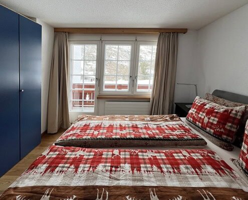 Gemütliches Schlafzimmer mit Doppelbett, rotem Karo-Bettwäsche, großen Fenstern und Blick auf die Natur, ideal für einen erholsamen Aufenthalt bei Matterhorn Home Apartments.