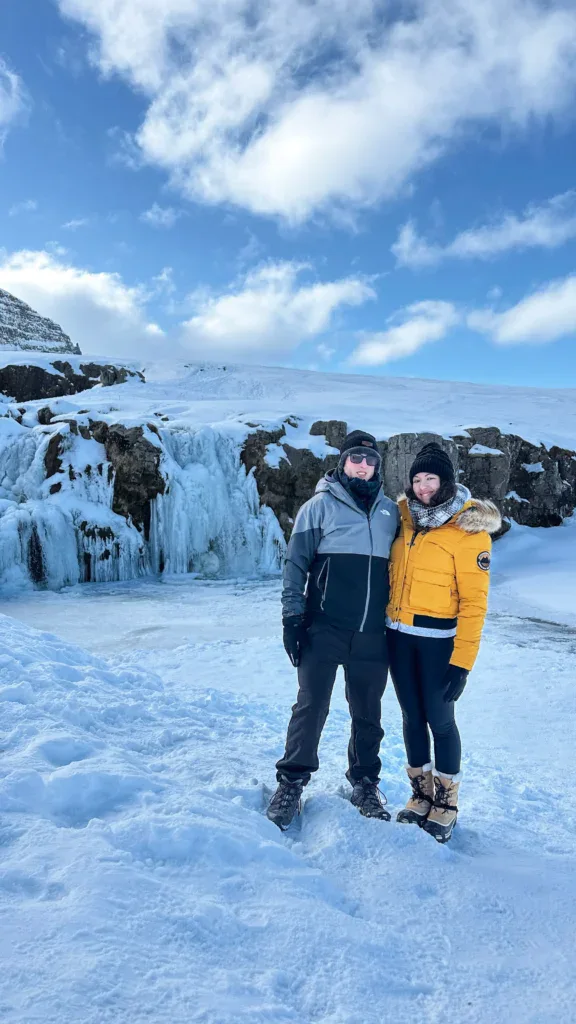 A couple dressed in winter gear stands on a snowy landscape in front of a frozen waterfall near Kirkjufell, Iceland. The man wears a black and gray jacket with snow pants and boots, while the woman wears a bright yellow winter coat with fur-lined boots. The sky is blue with fluffy clouds, and the icy terrain stretches behind them.