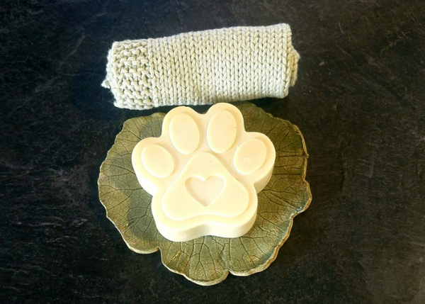 A cream grass-fed tallow paw print dog soap with a heart detail, resting on a decorative leaf dish, alongside a natural knitted wash cloth, handmade by Fatglow in Taranaki New Zealand
