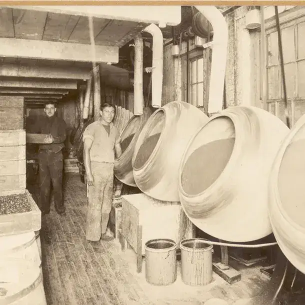  A black and white photo of two men in a factory standing beside a row of large, tilted, rotating industrial drums.