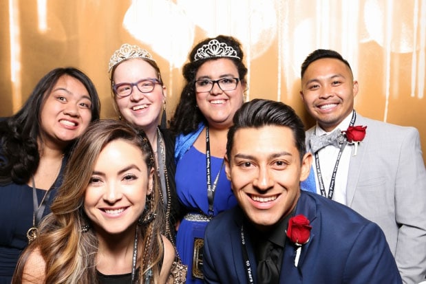 Group of six friends in formal attire posing for a photo booth shot against a shiny gold backdrop.