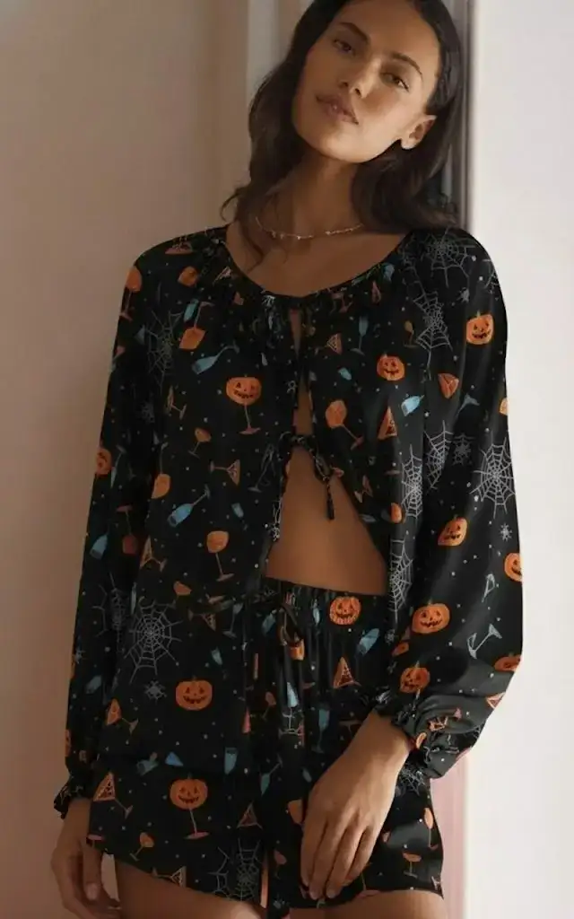 White long-sleeve top and ruffle shorts loungewear set decorated with cute black bows, bats, and orange pumpkins, contrasting orange cuffs, ideal for cozy fall mornings.