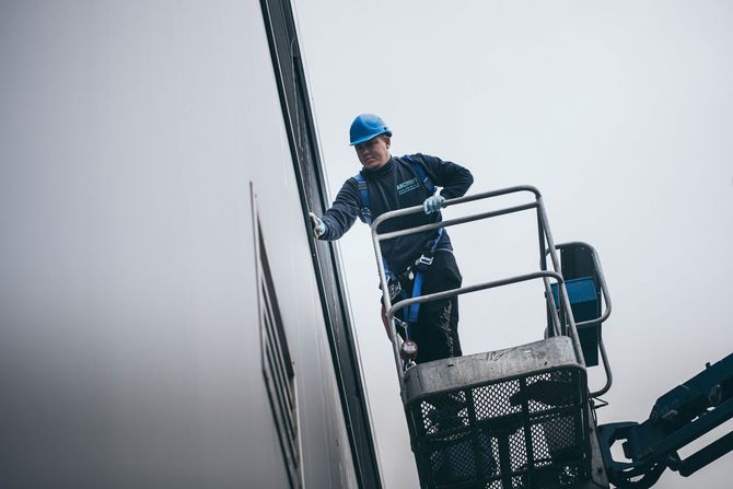 Ein Arbeiter mit blauem Schutzhelm und Sicherheitsgurt steht an einem nebligen Tag auf einer Hebebühne und arbeitet für eine Reinigungsfirma in Zürich an der Außenseite eines Gebäudes.