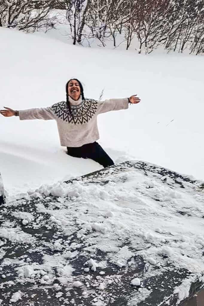 Person smiling and enjoying snow in a winter landscape with trees in the background.