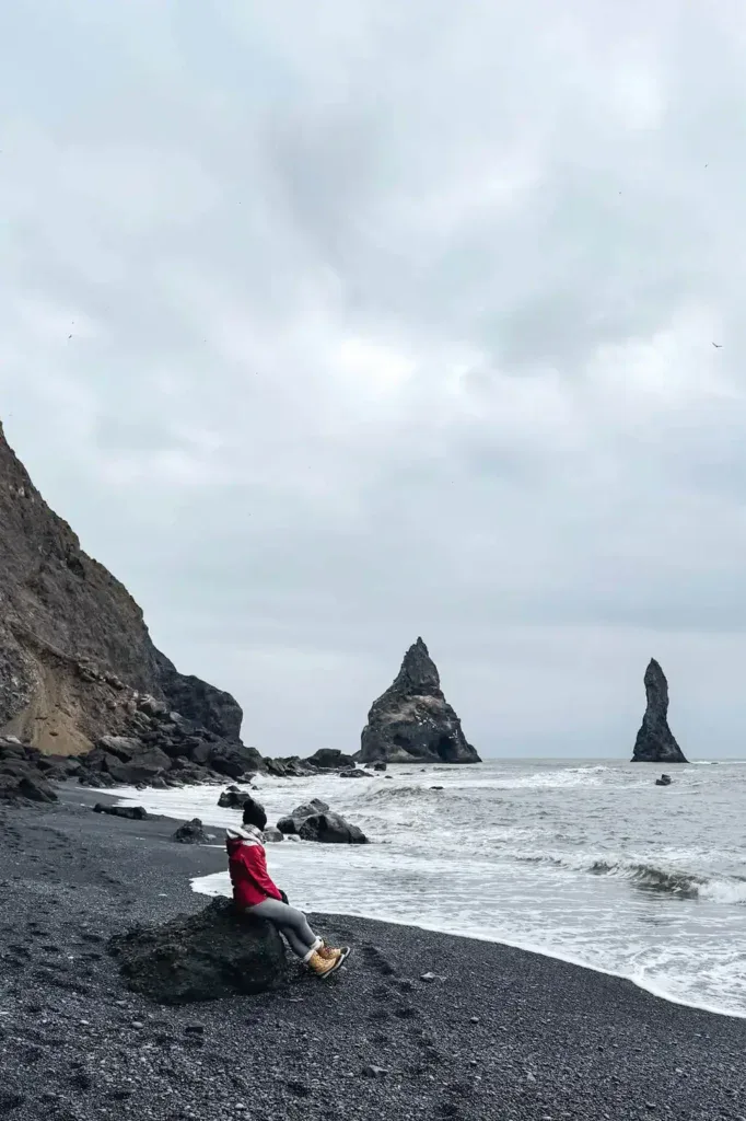 A person in a red jacket and beige boots sits on a black rock at a black sand beach in Iceland, gazing toward the ocean. In the background, dramatic basalt sea stacks rise from the water beneath an overcast sky, creating a striking and moody coastal scene. The rocky cliffside to the left adds depth to the rugged landscape