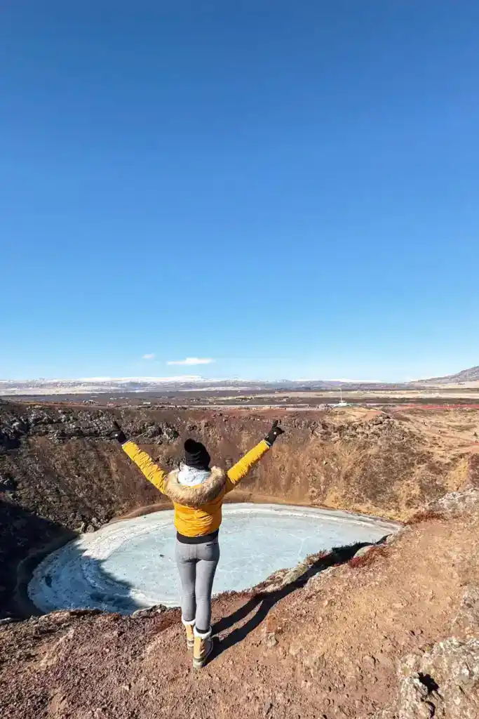 A person wearing a bright yellow jacket and winter boots stands at the edge of Kerið Crater in Iceland, raising their arms in excitement. Below, the volcanic crater lake is partially frozen, surrounded by reddish-brown slopes and rugged terrain. The clear blue sky and distant snow-capped mountains add to the dramatic and breathtaking landscape.