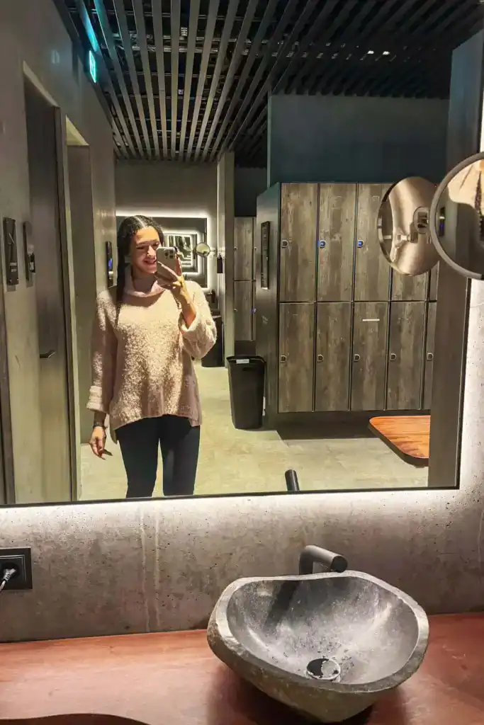 Woman taking a selfie in a modern bathroom with grey lockers and a stone sink.