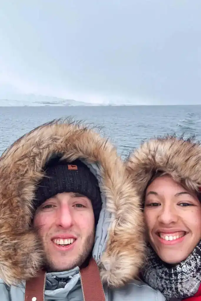 Two smiling people in winter coats with fur hoods taking a selfie by the sea in cold weather.