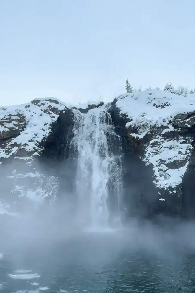 Snow-covered cliffs with a waterfall cascading into a misty pool below in a cold, wintry landscape.