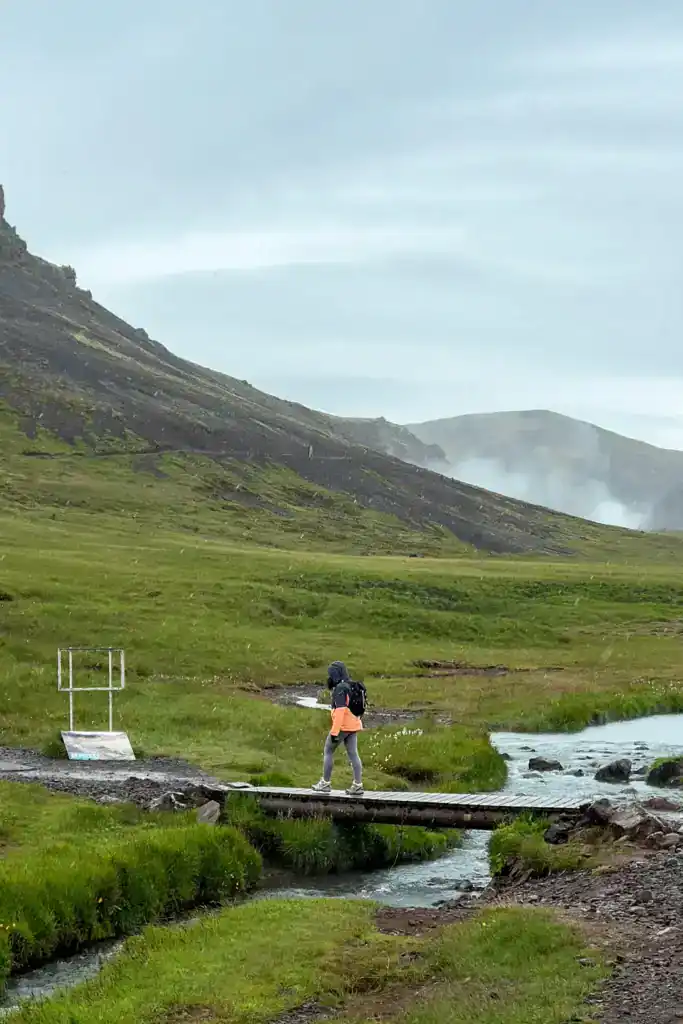 Hiker crossing a small wooden bridge over a stream in a lush green valley with mountains in the background.