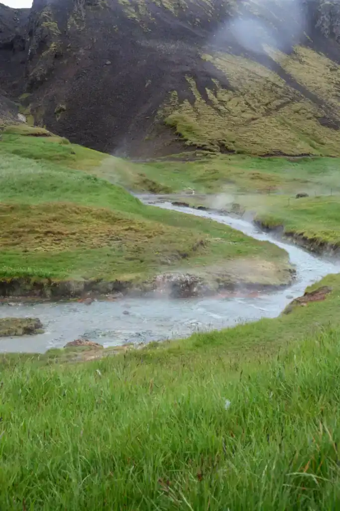 Steam rises from a hot spring river flowing through lush green fields in a mountainous landscape.