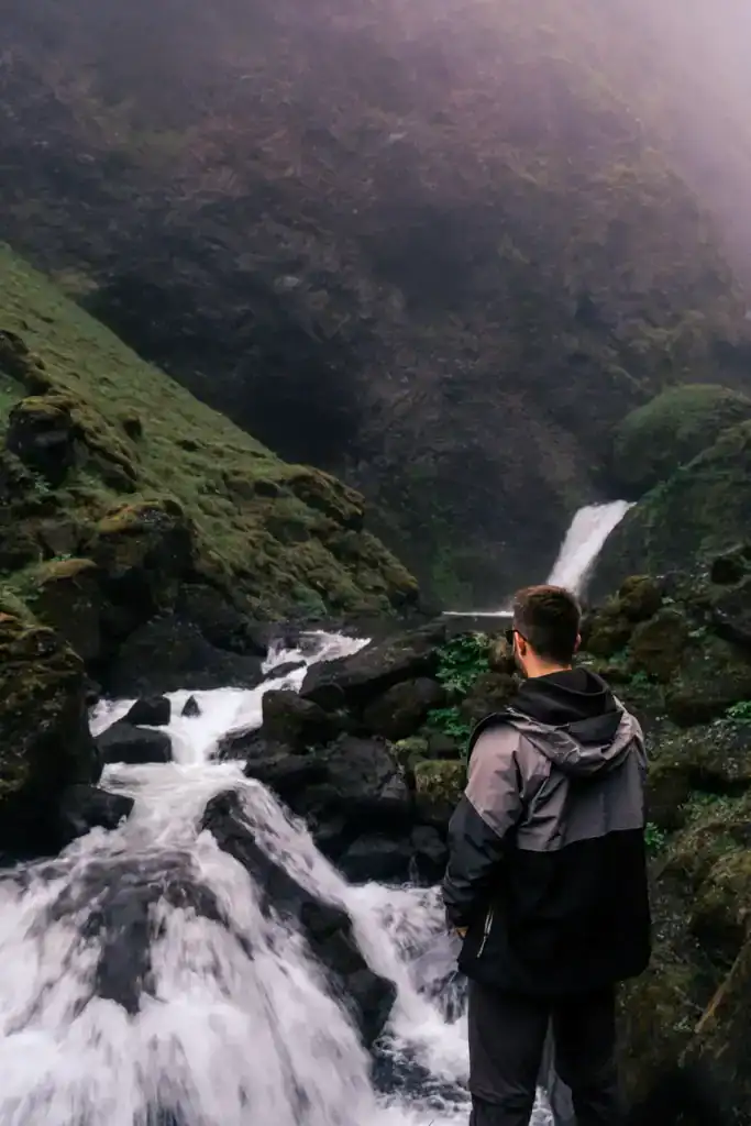 A person in a black and grey jacket stands by a rushing mountain stream with waterfalls in a lush green valley.