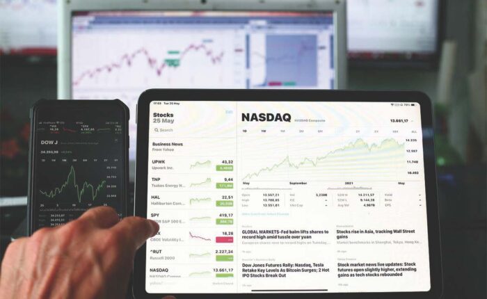 Hand zeigt auf Tablet mit NASDAQ-Chart; daneben Smartphone mit Dow Jones und Kursdaten.