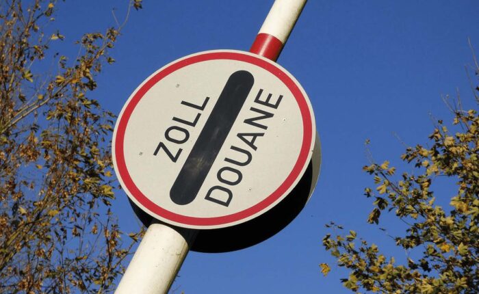 Rundes Verkehrszeichen mit der Aufschrift „Zoll/Douane“ vor blauem Himmel und herbstlichen Bäumen.