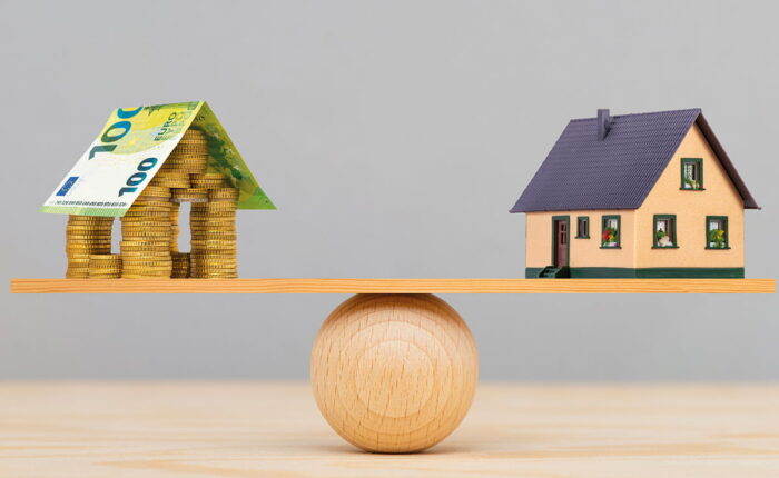 Haus aus Münzen und 100-Euro-Schein balanciert mit Modellhaus auf Holzbrett