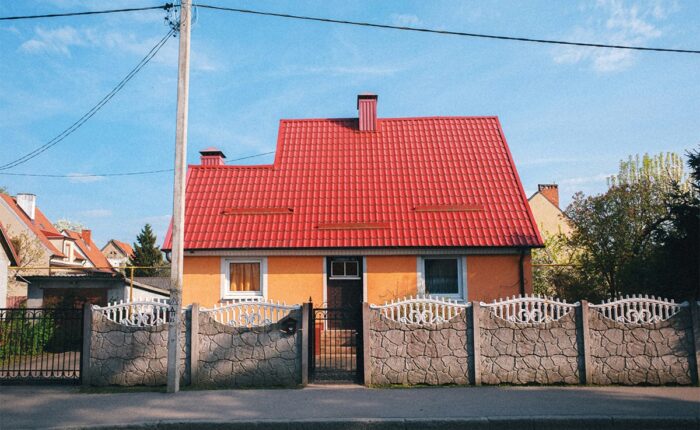 Einfamilienhaus mit rotem Ziegeldach und gelber Fassade hinter einem Steinzaun mit weißen Gittern.