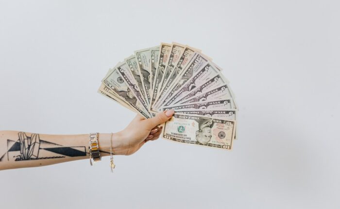 Eine tätowierte Hand hält eine Fächeranordnung von US-Dollar-Banknoten in verschiedenen Stückelungen vor einem weißen Hintergrund.
