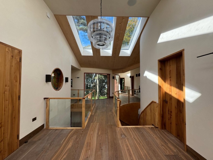 Modern upper-level hallway featuring warm wooden floors, Skylights, wooden doors, a glass balcony railing, and a central tiered crystal chandelier.