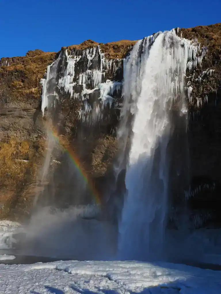 Frozen waterfall with ice formations and a rainbow at the base under a clear blue sky.