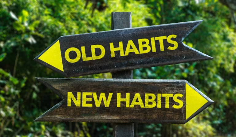 A wooden signpost for habit stacking with two yellow-on-black directional arrows pointing in opposite directions. The top arrow points left and reads "OLD HABITS." The bottom arrow points right and reads "NEW HABITS." The sign stands against a blurred background of green forest or jungle foliage, emphasizing a path forward.