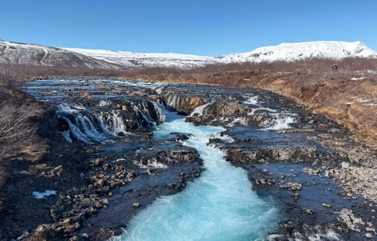 The stunning Brúarfoss waterfall in Iceland cascades over rugged volcanic rocks, with its signature icy blue waters flowing through a narrow gorge. Snow-capped mountains rise in the background under a clear blue sky, contrasting with the dark terrain. The vibrant turquoise water stands out against the rocky landscape, creating a mesmerizing natural scene.