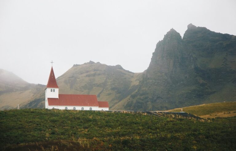 A white church with a red roof stands on a hill overlooking the town of Vík, Iceland. Below, the small coastal village is nestled between rugged cliffs and black sand beaches, with houses and buildings scattered across the landscape. In the distance, the Reynisdrangar sea stacks rise dramatically from the ocean as waves crash against the shore. The soft, golden light of the setting sun casts a warm glow over the town and surrounding mountains, creating a peaceful and picturesque scene.