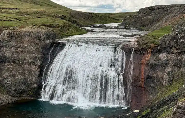 A wide waterfall cascading over a rocky ledge into a pool below, surrounded by green hills under a cloudy sky.