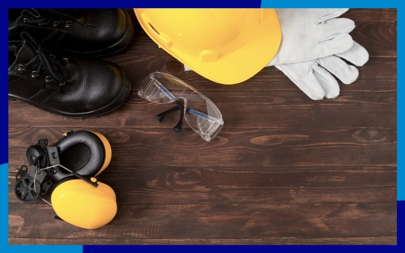 Banco di lavoro con sopra un casco giallo, guanti da lavoro, scarpe, occhiali e cuffie antirumore