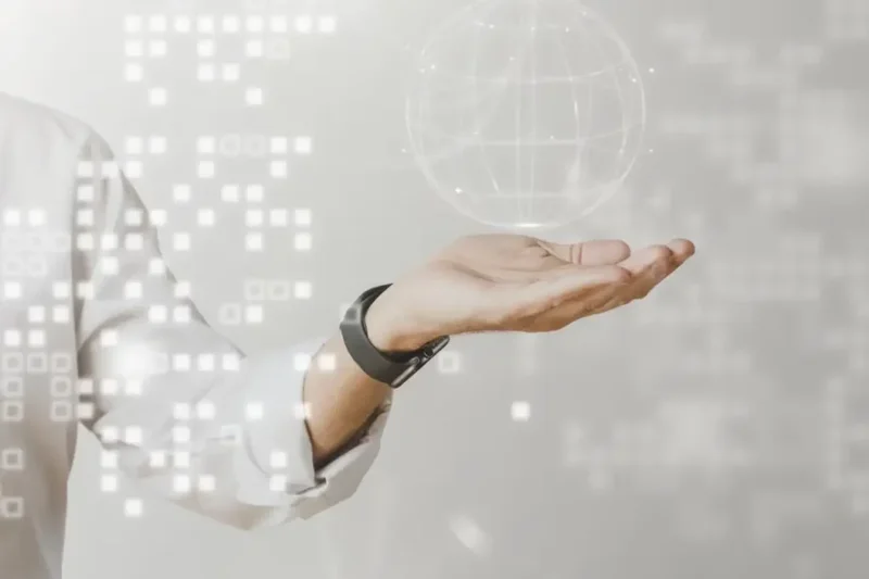 A person in a white shirt extends their hand upward. Above the hand, a digital sphere with interconnected lines and nodes hovers. White, pixelated squares surround the hand, suggesting technology or data transfer.