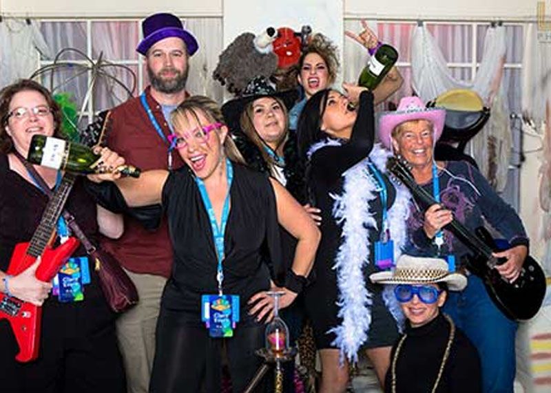 Group of partygoers in colorful outfits posing in a photo booth, wearing hats, boas, and props.
