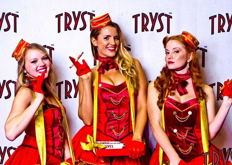 Three women in matching red carnival outfits with gold trim and feather headpieces pose on a backdrop with TRYST logos.