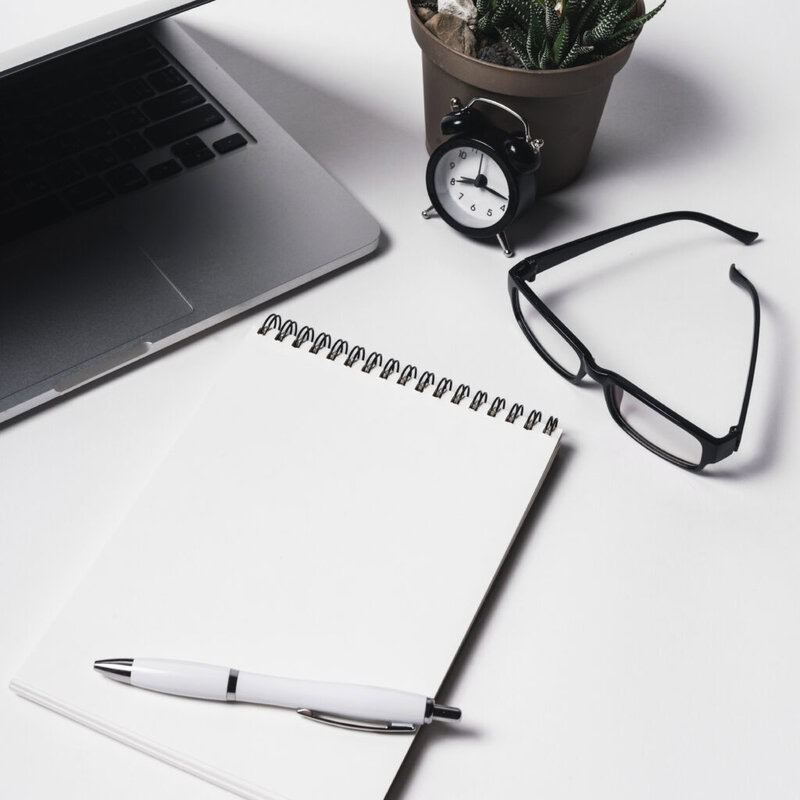 A workspace includes an open laptop, a blank spiral notebook, a pen, black glasses, and a small potted plant. An analog clock beside the plant shows approximately 10:10. The scene is set on a white surface.
