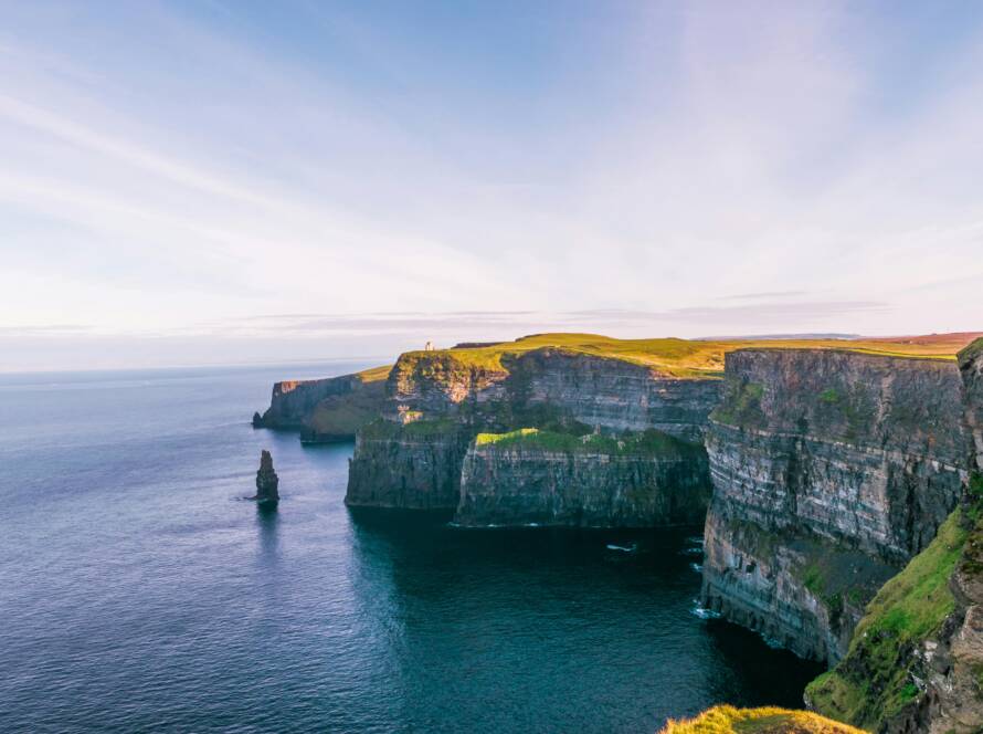 The image shows the dramatic Cliffs of Moher in Ireland, with steep rocky cliffs rising from the blue sea, topped with green grass, under a bright, partly cloudy sky.