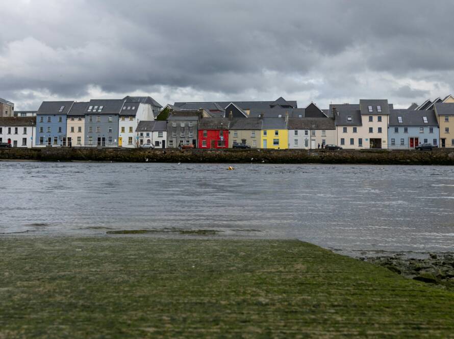 A row of colorful houses, including blue, red, and yellow, line the waterfront under a cloudy sky, with calm water in the foreground.
