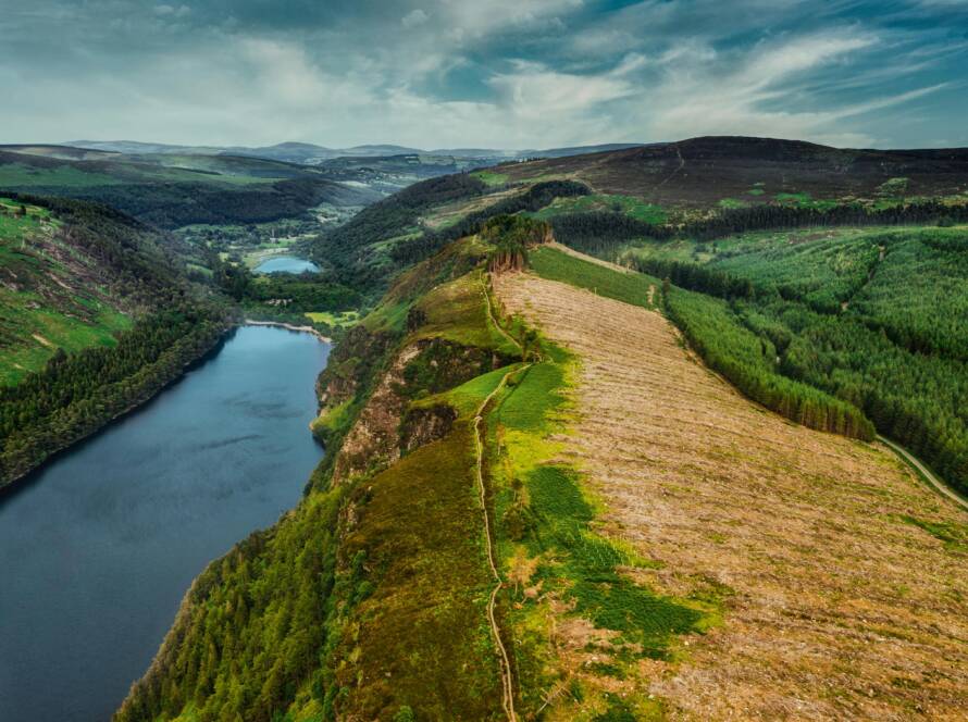 A winding river flows between lush green hills and a section of deforested land under a cloudy blue sky, with distant mountains on the horizon.