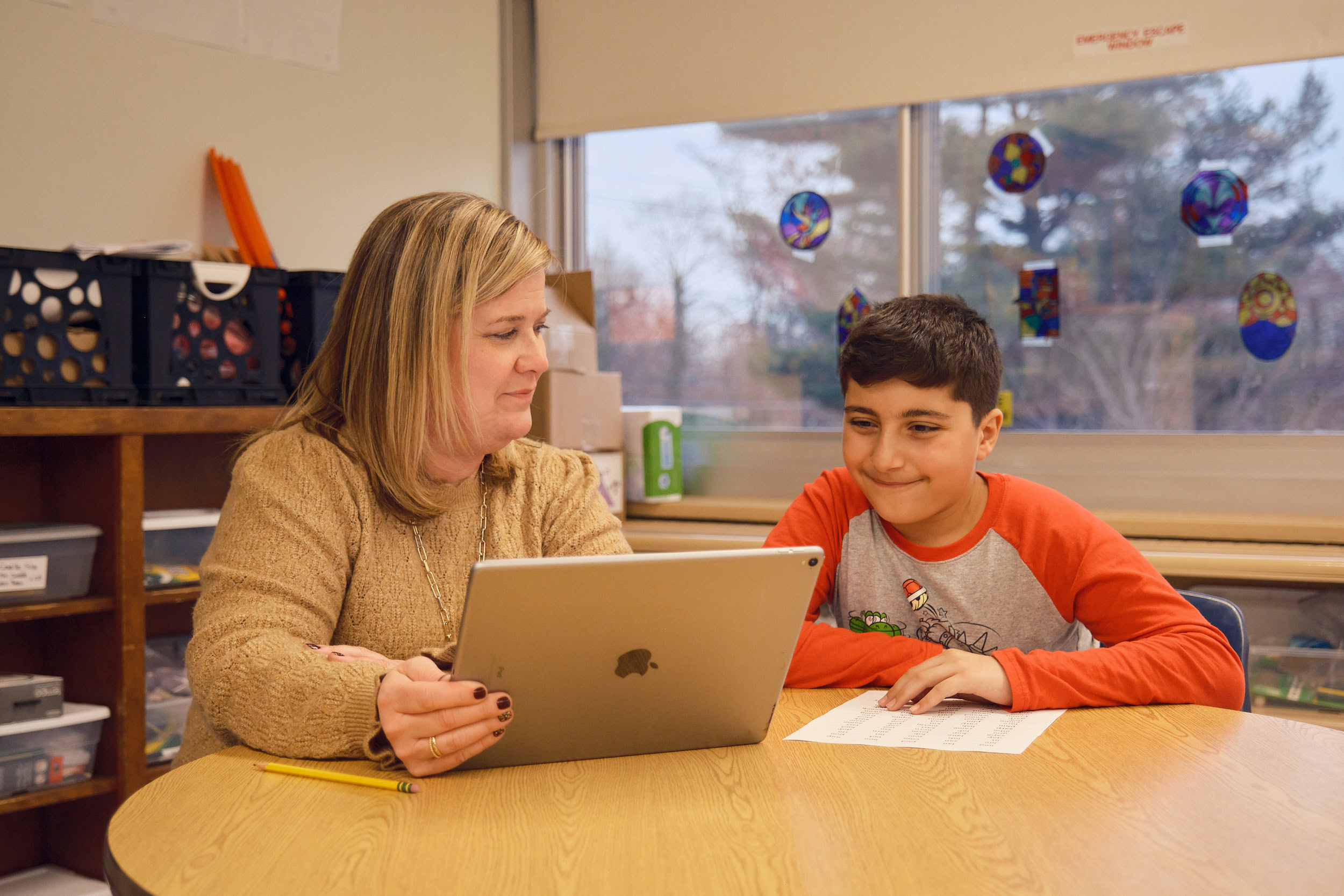 A woman and a boy sit at a table in a classroom, looking at a tablet together. The boy is holding a paper and smiling.