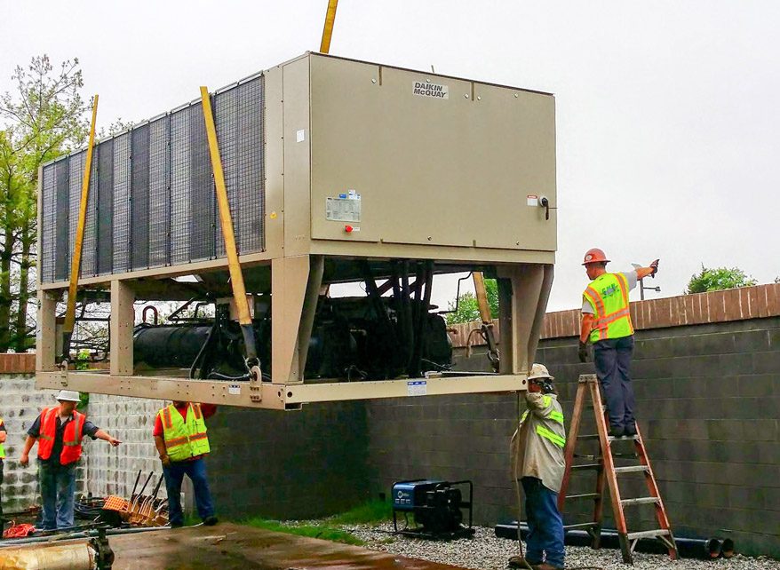Workers install a large industrial unit using cranes and ladders.