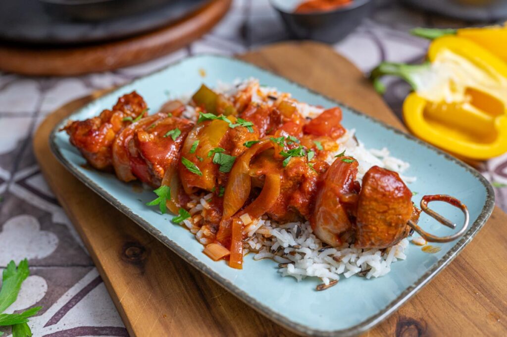 Nahaufnahme von Schaschlik-Spießen mit Schweinefleisch, buntem Gemüse und Zwiebeln in einer aromatischen Tomatensoße, serviert mit Reis. Ein klassisches Schaschlik Rezept für jeden Anlass.