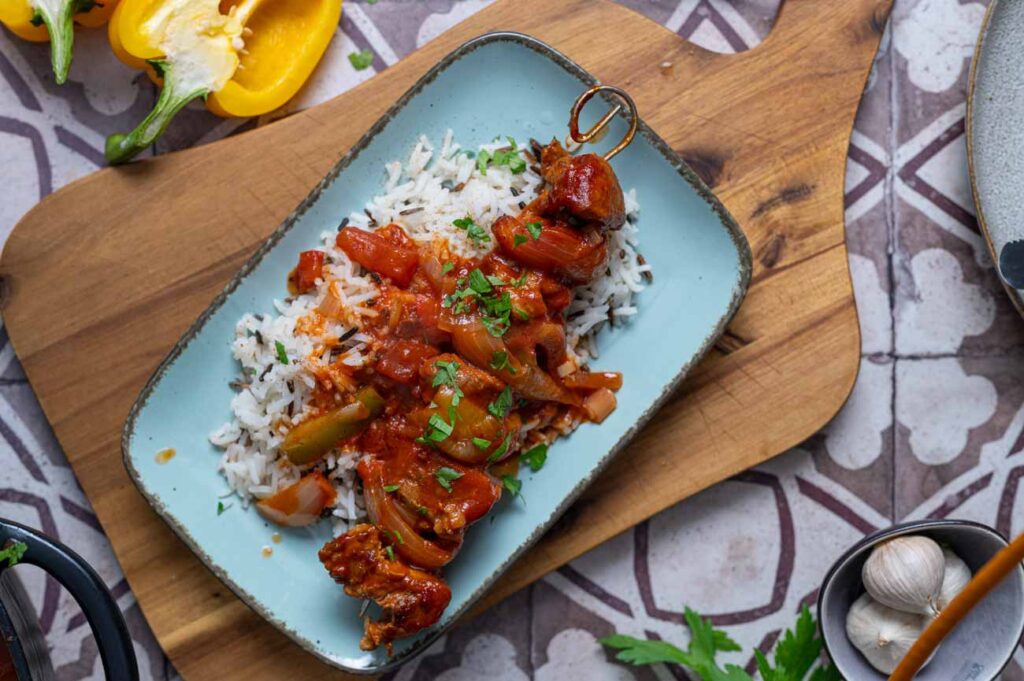 Schaschlik Spieße mit saftigem Schweinefleisch, Paprika und Zwiebeln, serviert in einer würzigen Tomatensoße. Im Hintergrund ein Teller mit dampfendem Reis – ein herzhaftes Schaschlik Rezept, perfekt für ein gemütliches Essen.