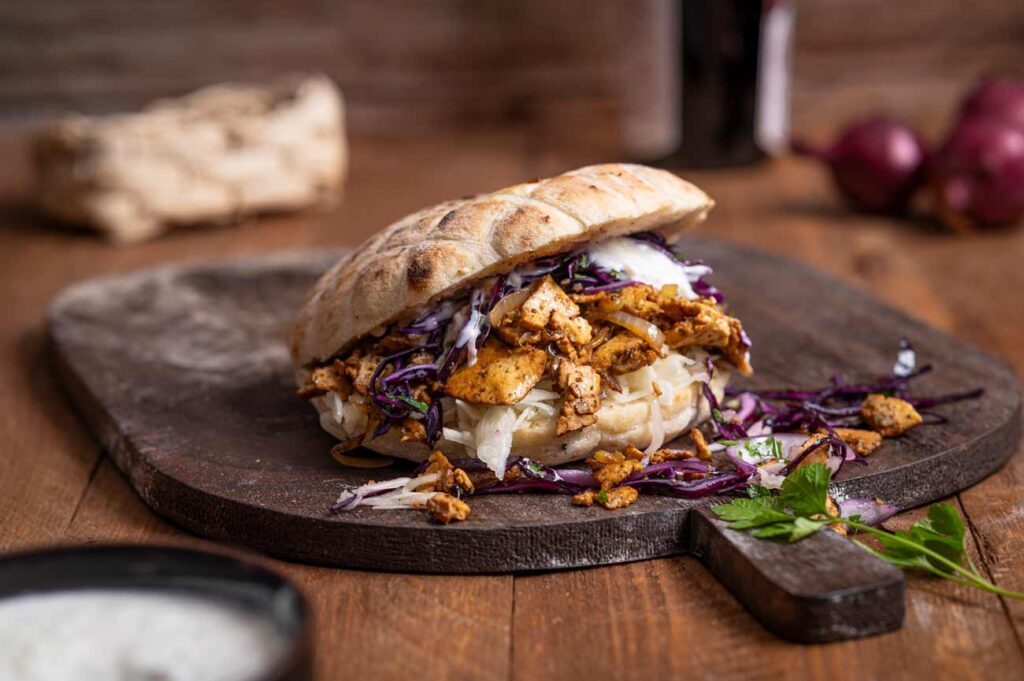 Frische Zutaten für einen veganer Döner, darunter gehobelter Räuchertofu, geschnittener Rotkohl, veganes Tzaziki und Fladenbrot.