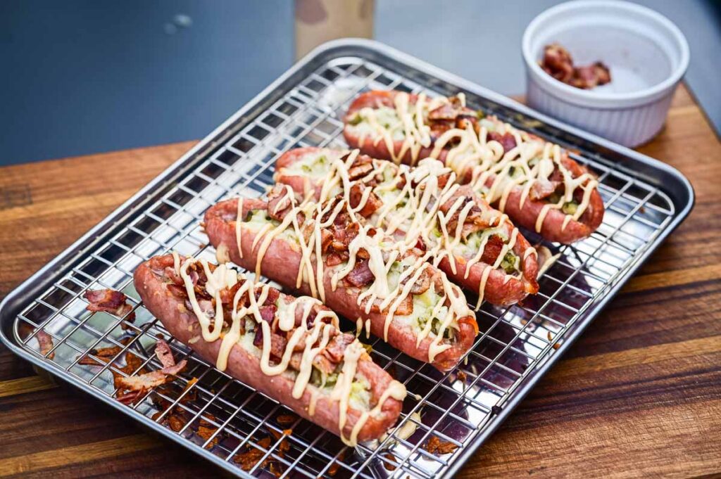 Heiße Bratwürste direkt vom Grill mit geschmolzener Käsefüllung, angerichtet mit Toppings auf einem Holzbrett