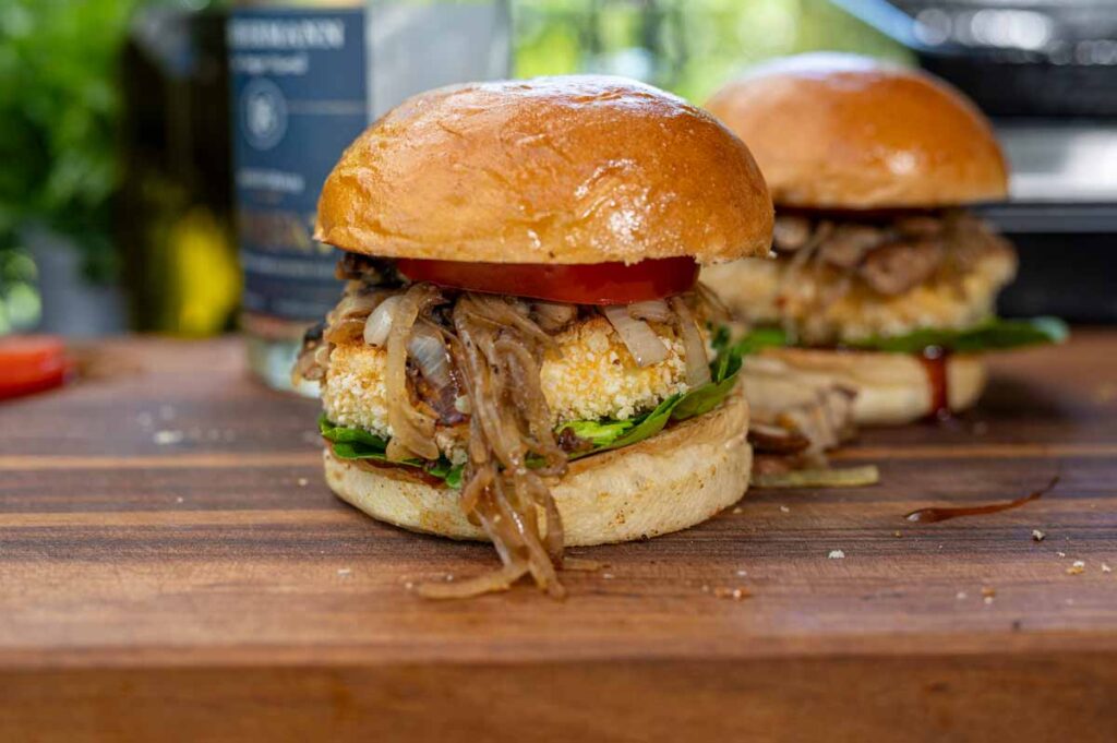 Angeschnittener vegetarischer Burger mit paniertem Grillkäse, der saftige Innenleben zeigt, umgeben von Salat und Tomate.