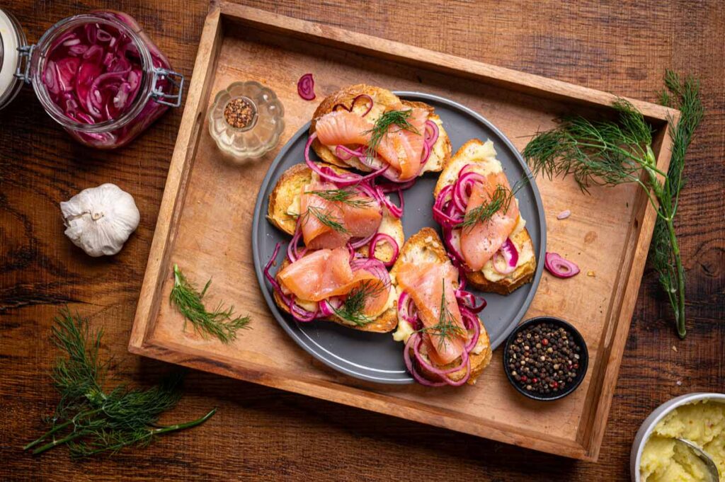 Fertige Crostini mit Lachs, cremiger Skordalia und eingelegten roten Zwiebeln auf einem Holzbrett serviert