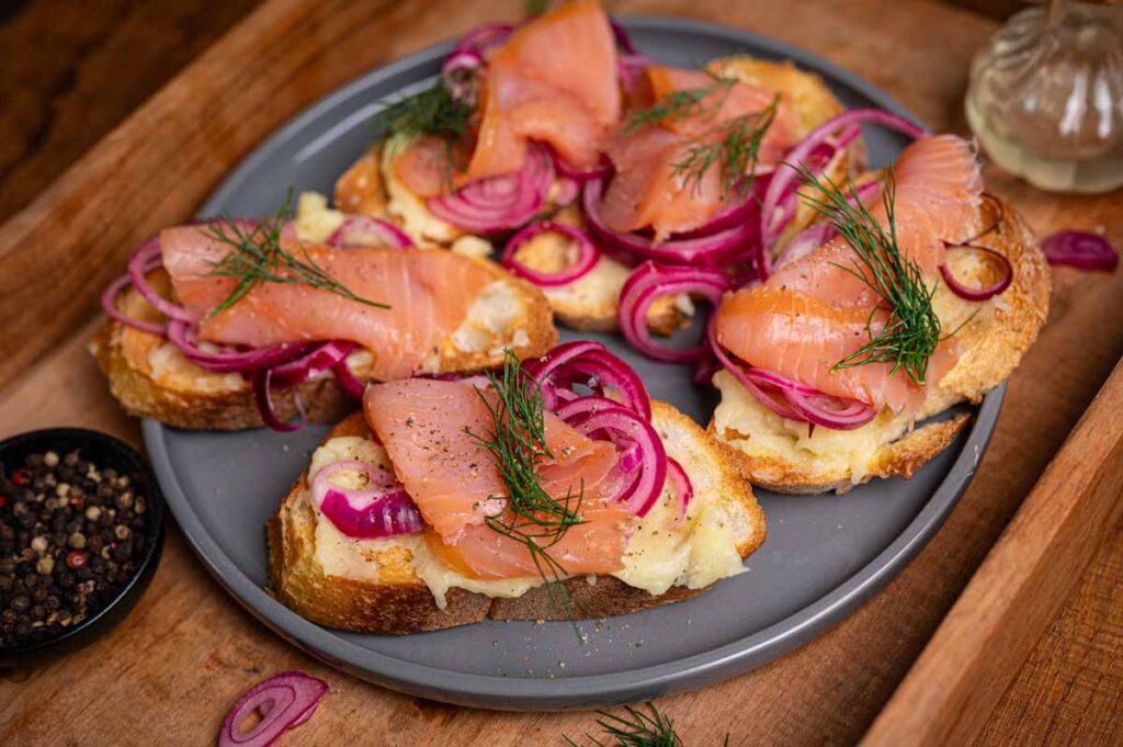 Gericht angerichtet auf einer Servierplatte – Crostini mit Lachsfilet, Kartoffelcreme und Zwiebelringen als Appetizer