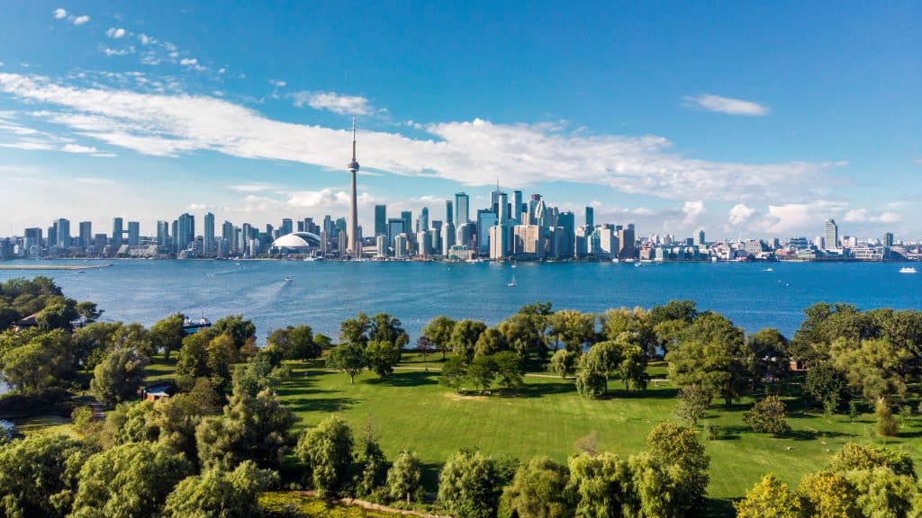 Aerial view of a coastal city skyline with tall buildings, green trees, a large harbor, and the blue ocean in the background.