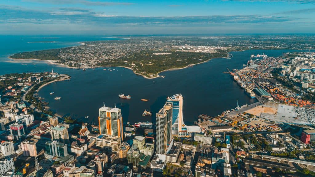 Aerial view of a coastal city skyline with tall buildings, green trees, a large harbor, and the blue ocean in the background.